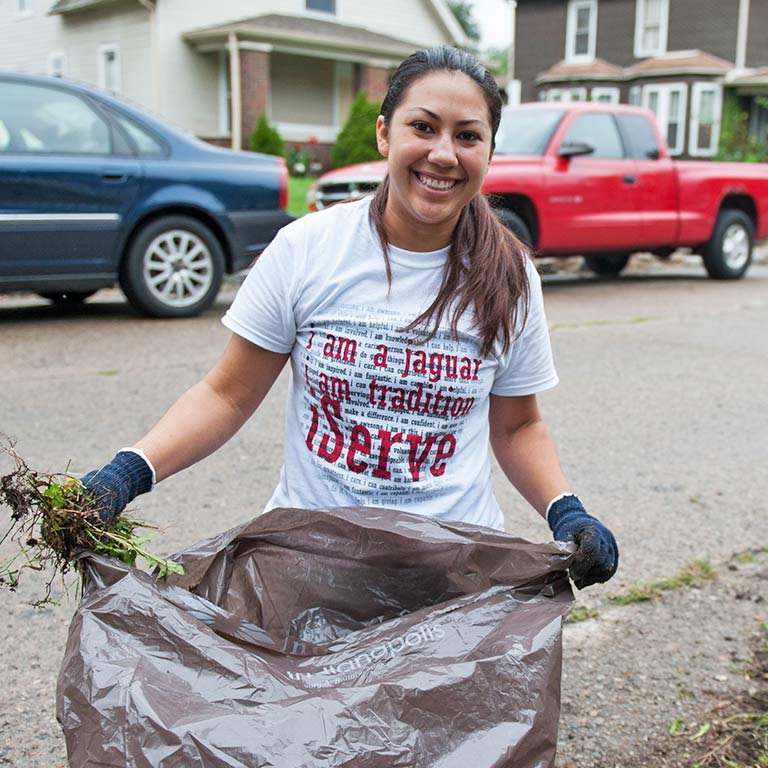 A student holds a large bag and a handful of yard waste. The text on her shirt says, 'I am a Jaguar, I am tradition, iServe.'