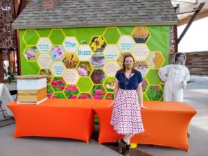 A person stands in front of an orange table with a bee hive and a model of a bee keeper in the background.