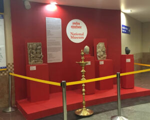 A small museum exhibit with red a wall, floor, and pedestals. a large white text panel is hanging on the wall next to a circular title. Four objects sit on pedestals in from of the texts. 