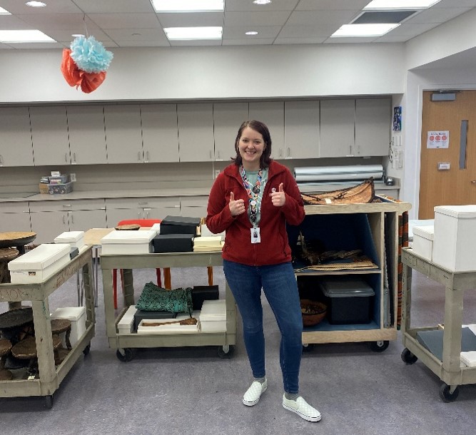 Katie is standing in front of four carts full of inventoried education collection objects with two thumbs up.