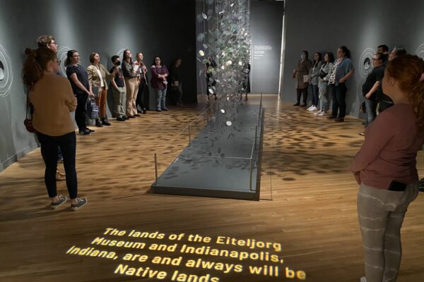 A group of people stand around the edges of a gallery with a sculpture in the middle of the room and text projected onto the floor: "The lands of the Eiteljorg Museum and Indianapolis, Indiana, are and always will be Native lands"