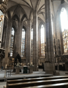 The image shows a church nave and altar, from St. Sebaldus Church in Nuremberg, Germany, one of the oldest buildings in the city. It combines Romanesque, Gothic, and Baroque elements into its design, here with the ribbed vaulting visible and several stained glass windows. Behind the altar is the Shrine of St. Sebaldus, which is unique for the marvelously sculpted metal snails upholding the structure. Both the shrine and the stained glass are mostly intact and original to the structure pre-WWII bombing. In this photo, light is coming in through a window on the right-hand side. Some pews are visible toward the bottom of the photo, and cropped from the left of the photo is an ornate pulpit, with a carving of a golden young child on the front, set against a greenish-blue background. The stonework used on the walls and ceiling is more warm in its gray hues, while in contrast, the floor has quite neutral and slightly cool greys for its stone. Statues and figures are between the stained glass windows, and just below them seems to be a series of painted reliefs depicting biblical scenes. The altar itself is a simple slab upon two heavy legs, and has plants with white flowers placed on top, alongside some candles, each in its own ornamental candle holder. A simple cross rests in the center of the altar, and in front of it, an open book. The photographer took the photo on the left-hand side of the room, creating a 3/4ths angle of the nave.